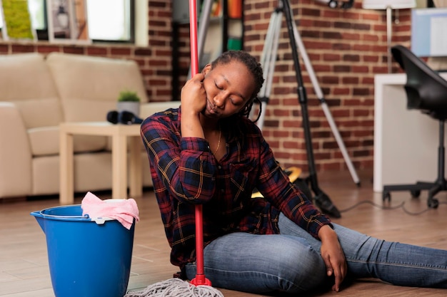 Exhausted housewife from daily work sitting on the room floor\
with no strength to continue cleaning apartment. overworked woman\
holding mop looks away with tiredness expression of\
exhaustion.