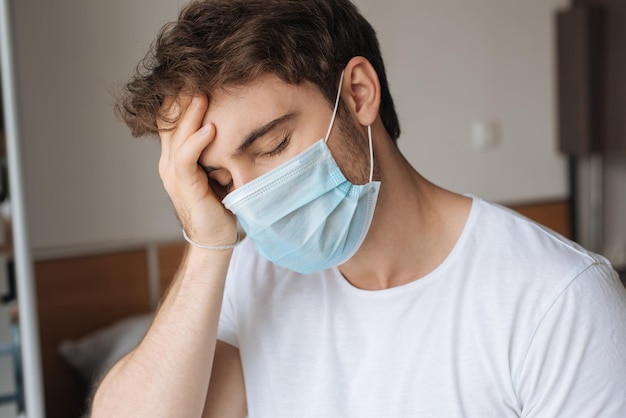 Exhausted ill man in medical mask sitting in bedroom during self isolation