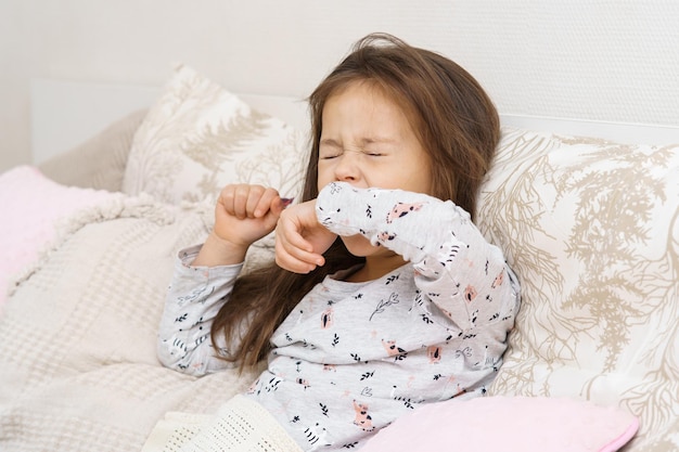Измученная маленькая длинноволосая брюнетка зевает или чихает с закрытыми глазами, прикрывая рот рукой в пижаме в постели