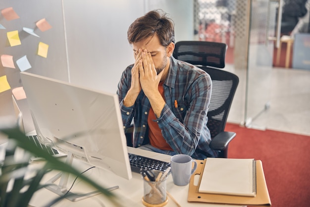 Exhausted male worker sitting at the table with computer and burying head in his hands