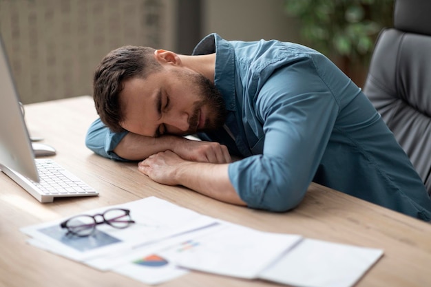 Exhausted man sleeping on desk in office next to laptop and documents