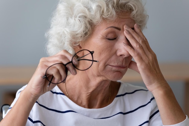 Exhausted mature woman take off glasses suffer from blurry vision or dizziness have high blood pressure tired senior female struggle with headache or migraine at home elderly sight problem concept