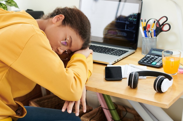 Exhausted mixed race teenager girl in glasses sleeping on desk with modern computer with stock market graphs on screen