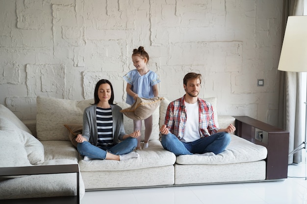 Exhausted mother and father sitting on couch feels annoyed tired while noisy little daughter shouting run around sofa.