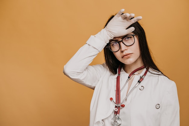 Exhausted nurse in glasses, medical gloves and phonendoscope on her neck, looks tired, touching her head.