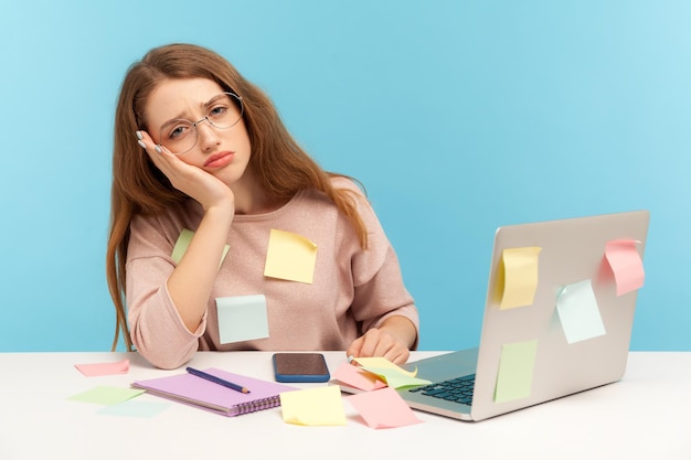 Foto donna esausta e oberata di lavoro con gli occhiali da nerd seduta coperta di note appiccicose, guardando con un'espressione annoiata e assonnata, stanca del carico di lavoro in ufficio a casa. tiro in studio indoor isolato su sfondo blu