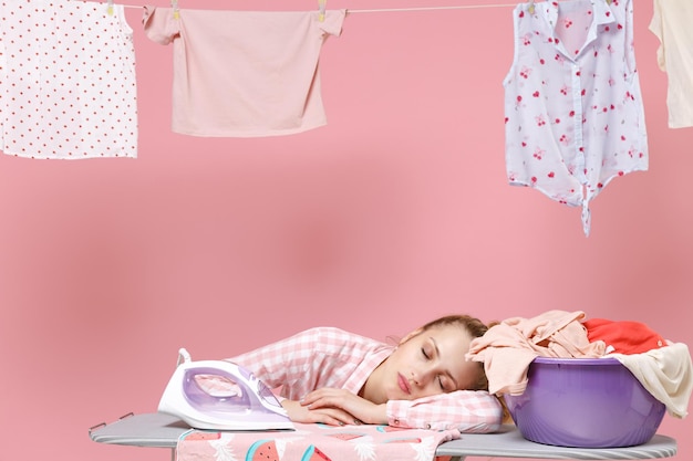 Exhausted tired young woman housewife in checkered shirt dry clothes on rope and ironing clean clothes while doing housework isolated on pink background studio. Housekeeping concept. Sleep on board.