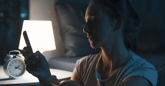 Photo exhausted woman sitting next to her bed and checking her smartphone late at night she is suffering from insomnia