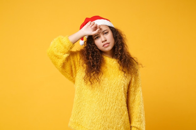 毛皮のセーター、黄色の壁の背景に分離されたクリスマス帽子で疲れ果てた若いアフリカ系アメリカ人サンタの女の子。明けましておめでとうございます 2020 お祝い休日のコンセプトです。コピー スペースをモックアップします。頭の上に手を置きます。