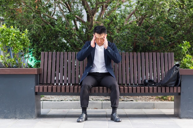 Exhausted young asian business man office worker sitting on bench in city park outdoors. stressed overworked male with a headache massages head. Depressed Tired employe suffering pain sick work