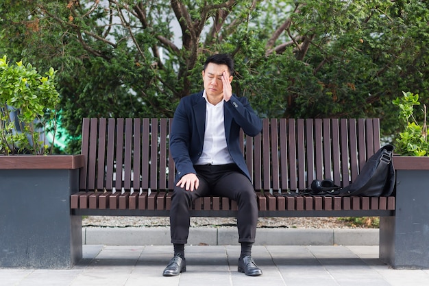 Exhausted young asian business man office worker sitting on bench in city park outdoors. stressed overworked male with a headache massages head. Depressed Tired employe suffering pain sick work