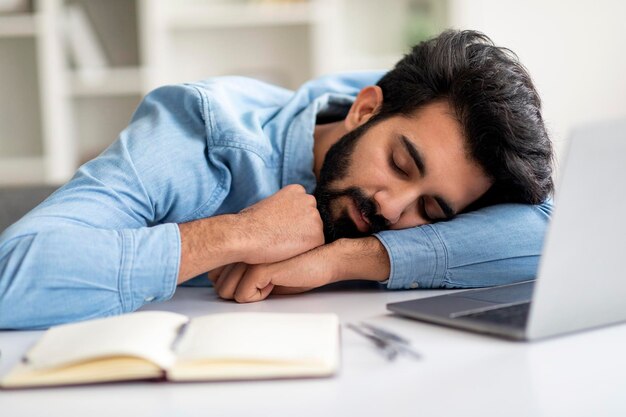 Exhausted young indian man sleeping at desk with laptop closeup