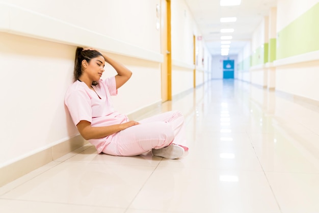 病院の廊下の壁に向かって床に座っている疲れ果てた若いラテン系看護師