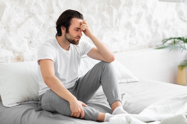 Exhausted young man sitting on bed touching his head