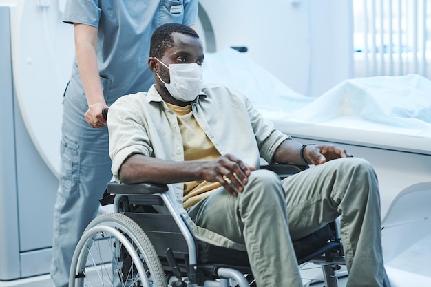 Exhauster black coronavirus patient in facial mask sitting on wheelchair pushed by nurse after MRI