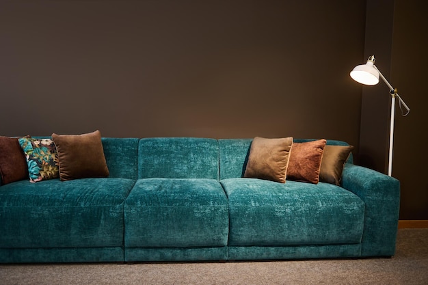 Exhibition of modern stylish upholstered furniture in the showroom of a furniture store Focus on a turquoise soft velour setee and brown pillows lit by a lamp against a brown wall background