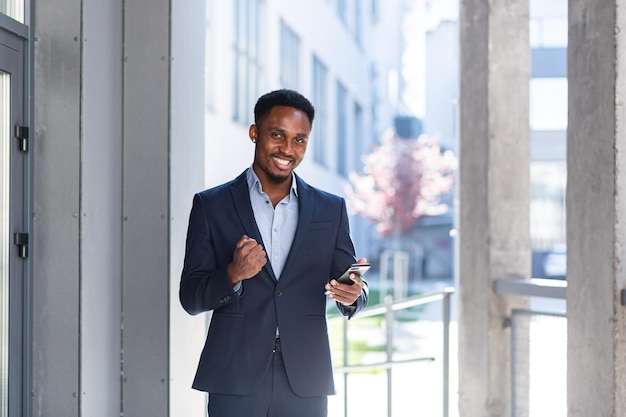 Exited African American Businessman Celebrating Achievement looking at Smartphone. Successful business man victory win with mobile phone outside. Happy Good News Success Deal, Startup Bet winning rate