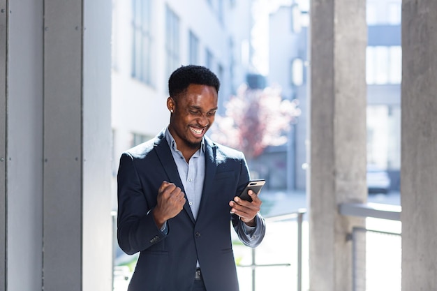 Uscito dall'uomo d'affari afroamericano che celebra il successo guardando lo smartphone. vittoria di successo dell'uomo d'affari con il telefono cellulare all'esterno. happy good news successo affare, tasso di vincita della scommessa iniziale