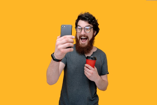 Exited bearded guy holding is taking a photo with smartphone and holding a cup coffee