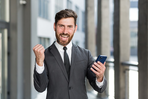 Uomo d'affari uscito che celebra il successo guardando lo smartphone all'aperto. uomo d'affari di successo che celebra la vittoria con il telefono cellulare all'esterno. buone notizie felici. affare di successo, scommessa di avvio, commercio