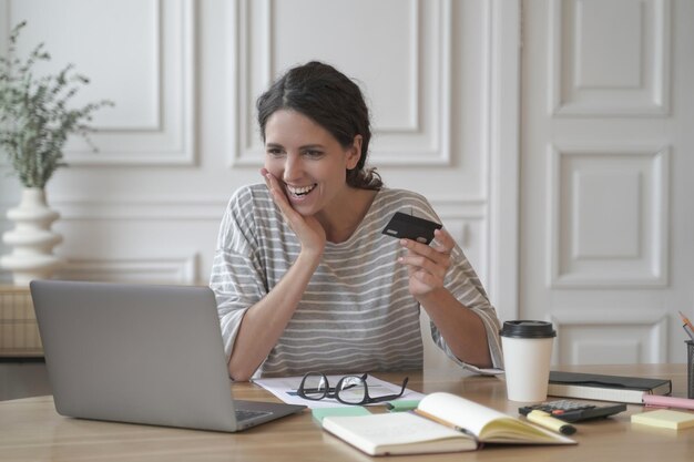 Exited italian entrepreneur lady joyfully looking at computer screen holding credit card