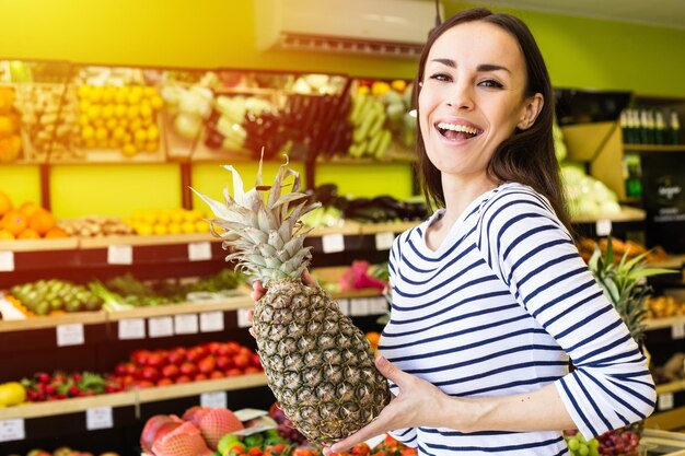 Exotic fruit for health an attractive smiling woman in casual clothes holds in her hands two large pineapples in the background of the shelves of the store of organic products