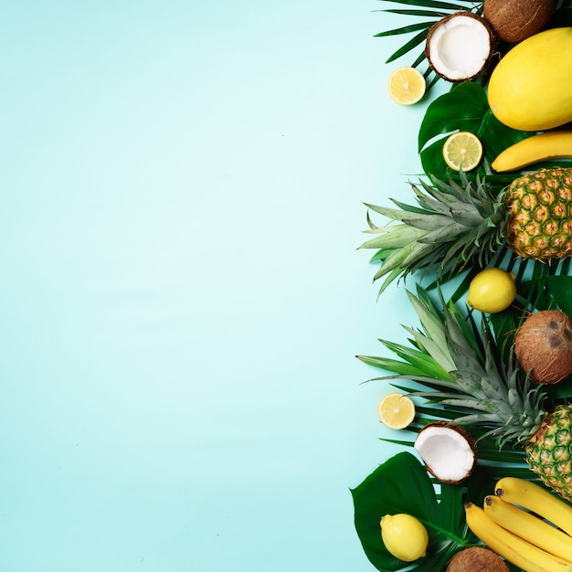 Exotic pineapples, ripe coconuts, banana, melon, lemon, tropical palm and monstera leaves on blue background