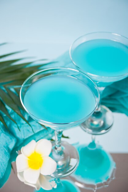 Exotic tropical Blue Curacao cocktail drink in a glasses with Plumeria frangipani flower, palm leaf on the surface.