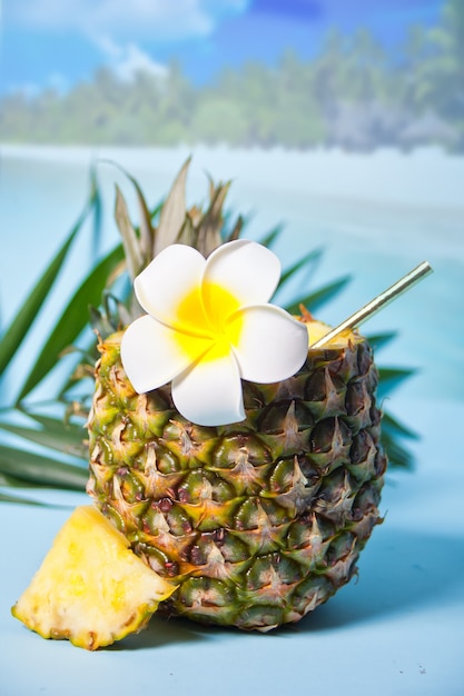 Экзотический тропический коктейль в ананасе с цветком франжипани Plumeria и пальмовым листом для украшения. Морское побережье на заднем плане.