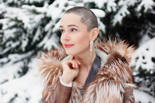 Exotische vrouw met kort haar, portret van de schoonheidsstijl. lachende, schattige, peinzende kale vrouw in de winter tegen de achtergrond van sneeuw en lange besneeuwde sparren. vrouw poseren in avondjurk