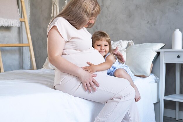 Photo expectant mother sitting on bed with little baby daughter stroking belly