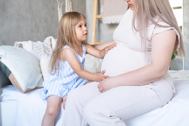 小さな赤ん坊の娘が腹をなでてベッドに座っている妊婦