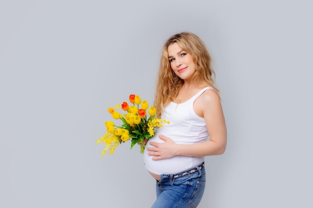 Expectant Mother Touches Her Stomach holding A bouquet Of spring flowers On A White Background