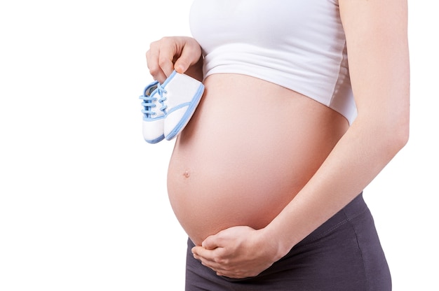 Ожидание. Обрезанное изображение беременной женщины, держащей маленькую обувь, стоя изолированной на белом