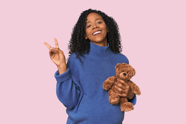 Ожидающий афроамериканца с плюшевым медведем радостный и беззаботный показывающий символ мира пальцами
