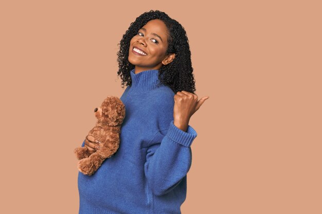 Photo expecting african american with teddy bear points with thumb finger away laughing and carefree