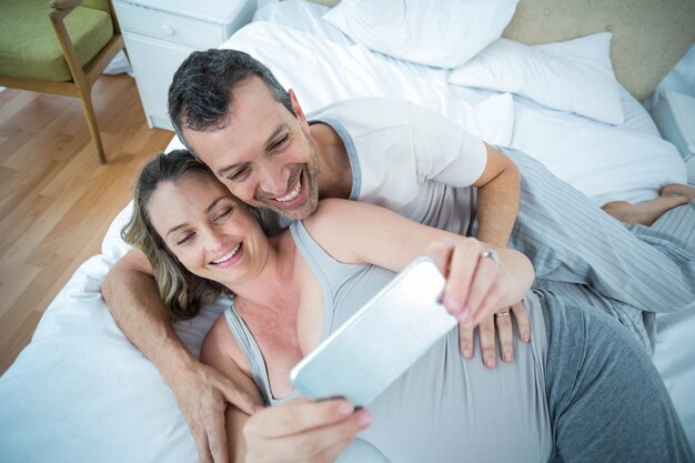 Expecting couple lying on bed and using digital tablet