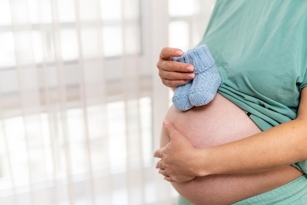 Expecting woman standing at the window and holding baby socks on her tummy Lady holding her one hand on the belly and covering her bellybutton