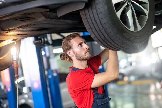 Experienced attentive young adult man doing auto repair looking at wheel at service station