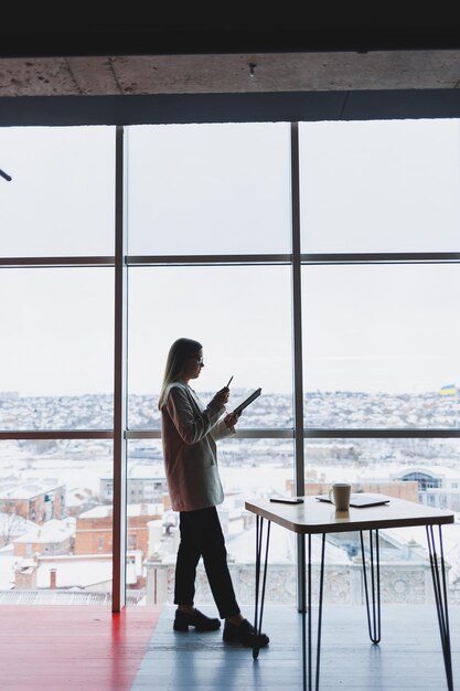 Experienced business woman holding a phone while standing in a modern office interior near a large window overlooking the cityscape Female executive looks pleased after a successful meeting