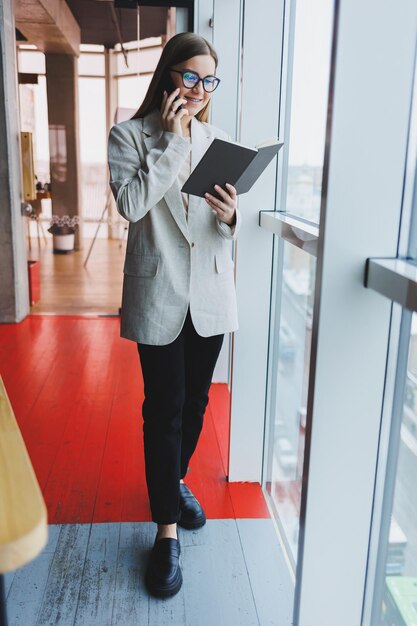 Experienced business woman holding a phone while standing in a modern office interior near a large window overlooking the cityscape Female executive looks pleased after a successful meeting