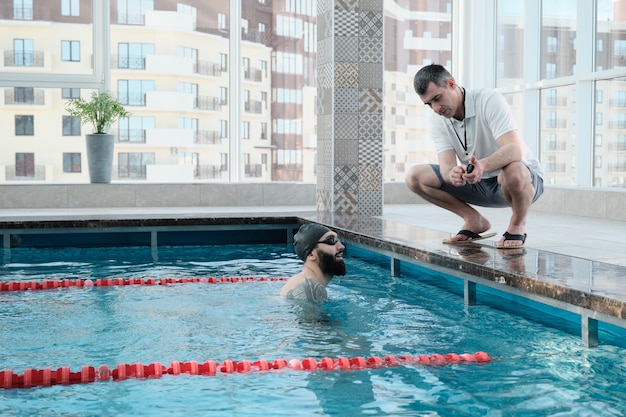 Experienced coach crouching on edge of poolside and showing time on stopwatch to swimmer while discussing his results