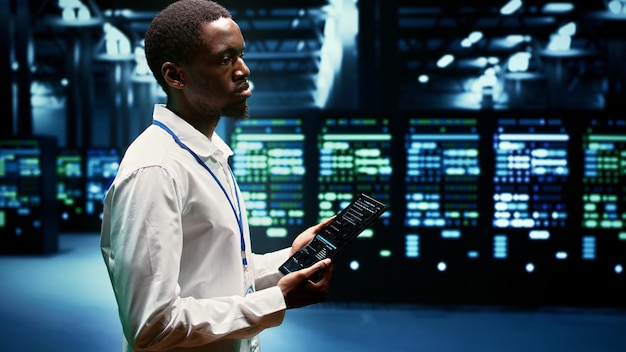 Experienced computer scientist maintenancing server rows delivering web content to online clients. African american supervisor using tablet to ensure critical systems functionality