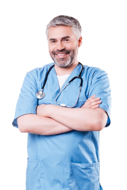 Experienced and confident surgeon. Cheerful mature doctor looking at camera and smiling while keeping arms crossed and standing isolated on white