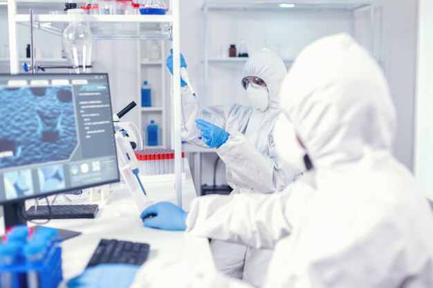 Experienced doctor taking sample from test tube using micropipette wearing sterile suit. Team of microbiologists in research laboratory conducting experiment during global pandemic with covid19.