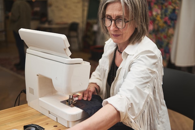 Experienced elderly female fashion designer tailor sewing clothes on sewing machine in a tailoring atelier