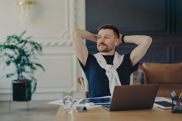 Experienced European man rests on chair looks away with thoughtful expression poses at desktop poses at home office listens information via earphones uses free internet connection prepares for exam