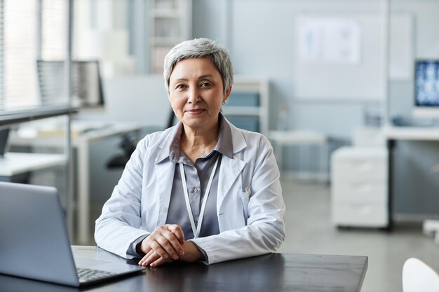 Experienced female doctor in lab coat sitting in front of laptop by workplace