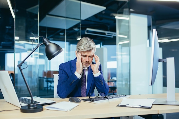 Foto un esperto uomo d'affari dai capelli grigi lavora al computer fino a quando la stanchezza tarda ha un forte mal di testa, stress e depressione