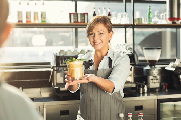 Experienced middle aged barista giving tasty cold coffee non-alcoholic cocktail to customer. Bartender at her working place. Small business concept, copy space
